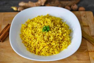 Primer plano de un tazón blanco con arroz basmati amarillo, sazonado con cúrcuma y decorado con un puñado de guisantes verdes en el centro. El plato está sobre una tabla de cortar de madera, con ramas de canela y jengibre fresco en el fondo.