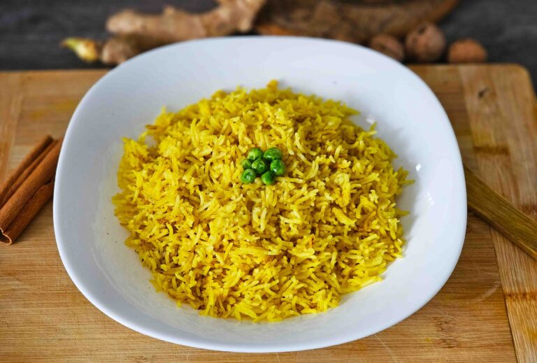 Primer plano de un tazón blanco con arroz basmati amarillo, sazonado con cúrcuma y decorado con un puñado de guisantes verdes en el centro. El plato está sobre una tabla de cortar de madera, con ramas de canela y jengibre fresco en el fondo.